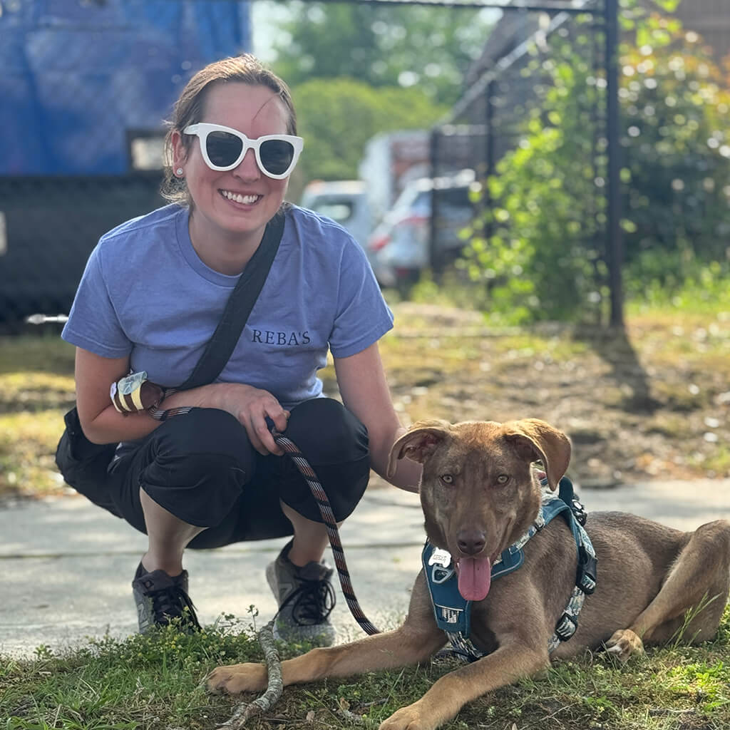 Volunteer or staff squats down next to brown dog. Dog is laying on the ground.