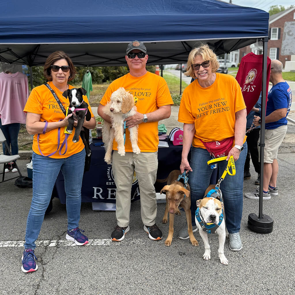 Three volunteers in orange shirts standing in front of Rebas booth. Two are holding dogs and the other lady has two dogs on leashes.