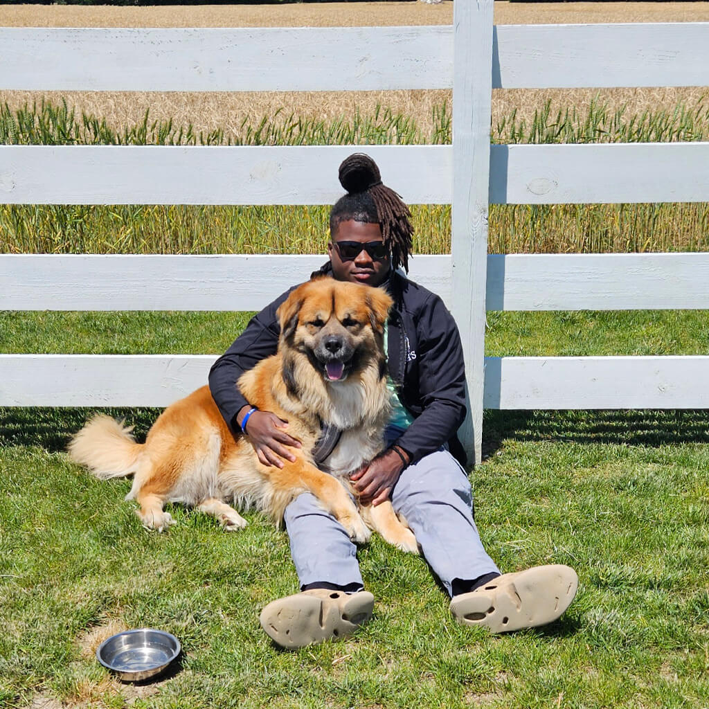 A staff member is resting against a white fence while holding the brown dog in his lap. There is a silver water bowl in the front.