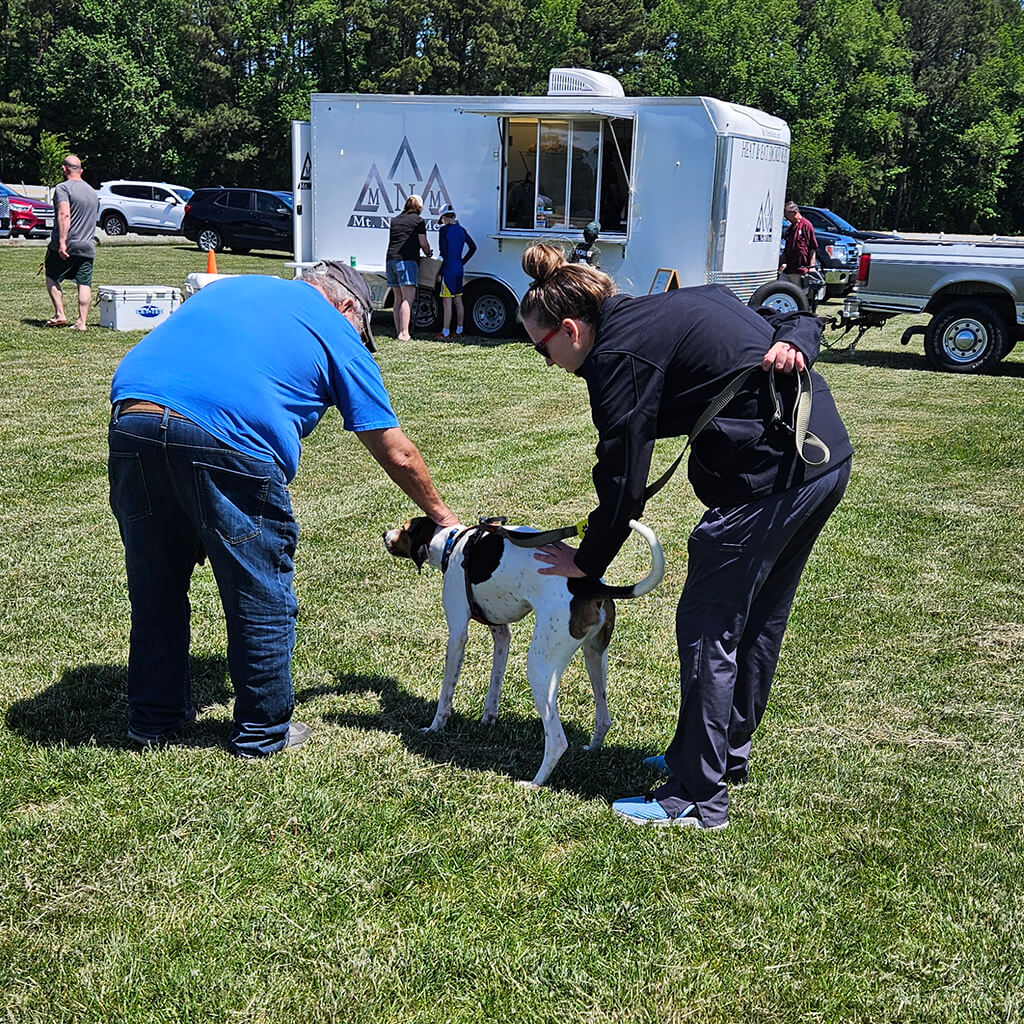 A volunteer is showing the tan and white dog to a visitor. They are both petting the dog. They are in a field with a white food truck in the background.