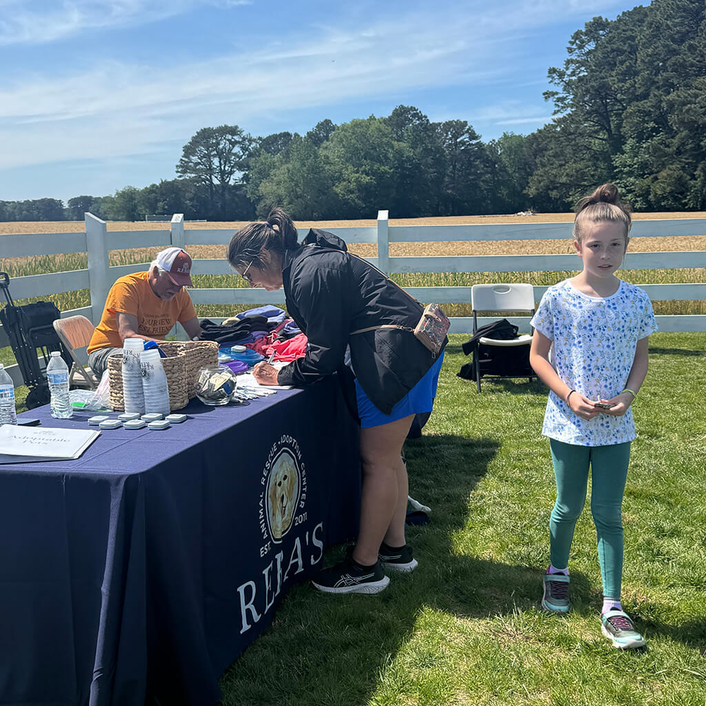 Reba's table with a visitor filling out a form, a volunteer behind the table in an orange shirt, and a little girl walking in front of the booth. The table is set up near a white fence in the farm field.