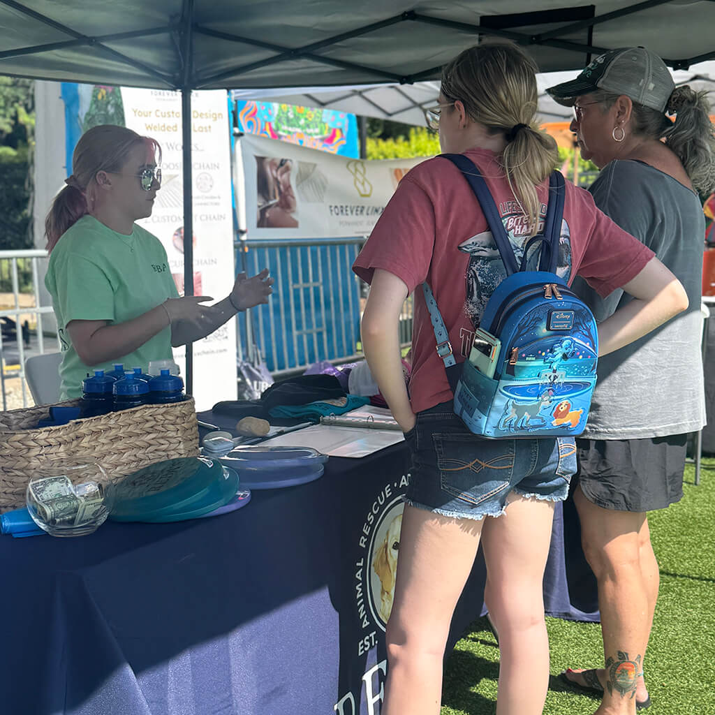Two visitors are visiting Reba's tent and talking to a volunteer in green behind the table with a blue table cloth.