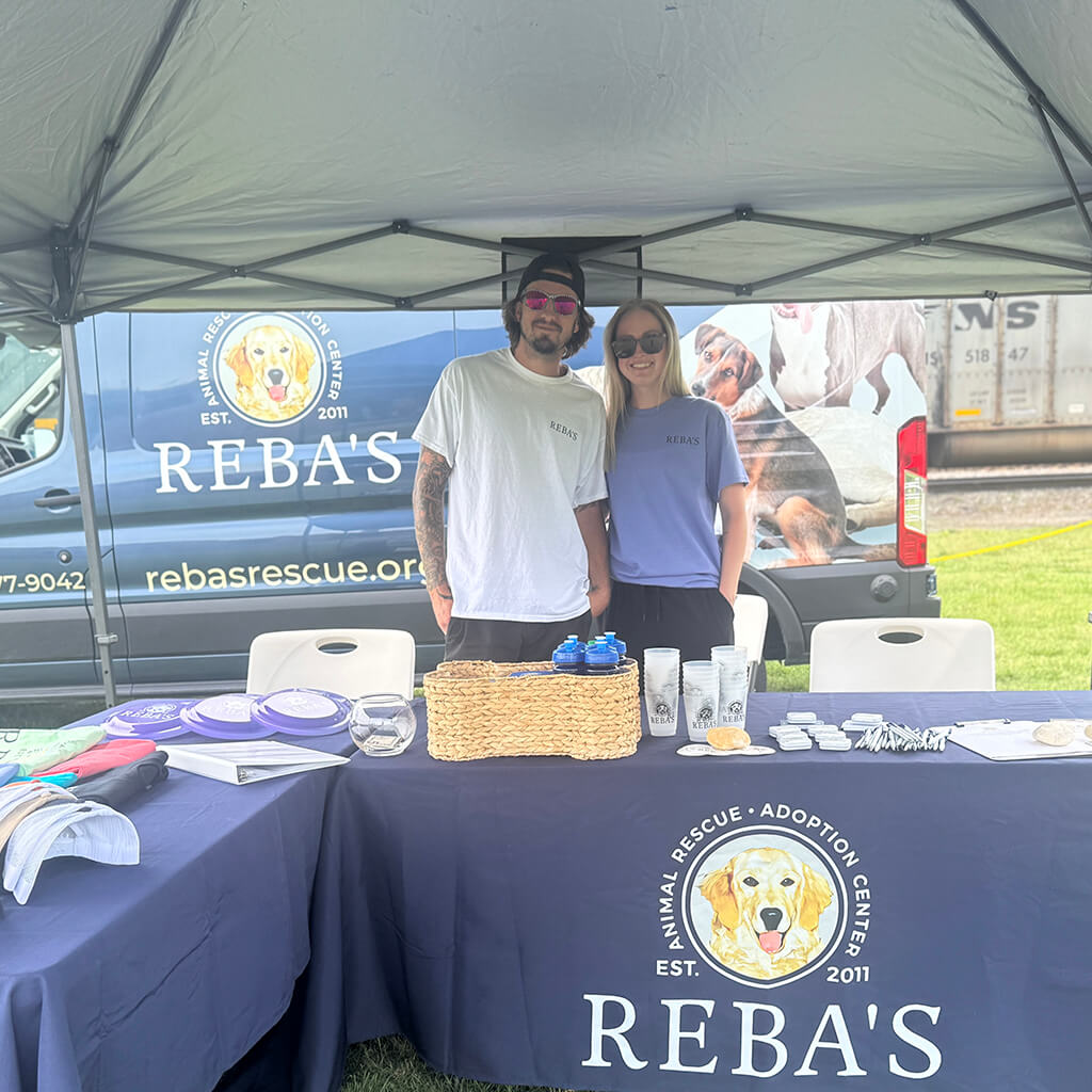 two staff members behind the Reba's table under the tent. The van is in the background and there is literature and Reba's merch on the table.