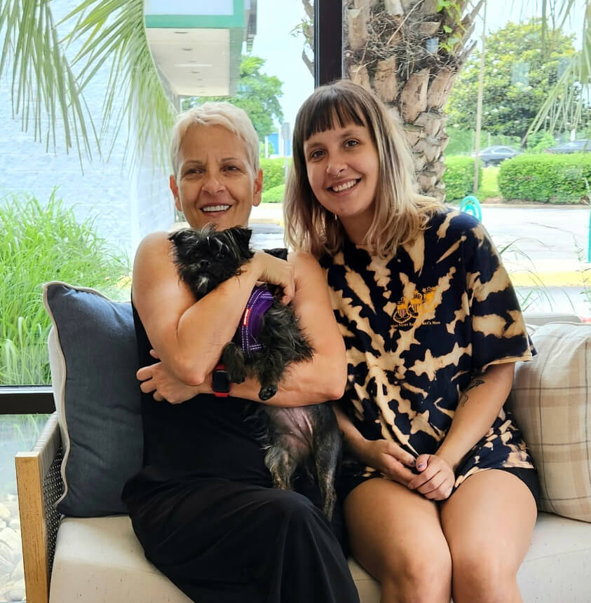 Two ladies that adopted Tippy. One lady is holding the black dog in her arms and the other lady is leaning in. They are posing in Reba's lobby area.