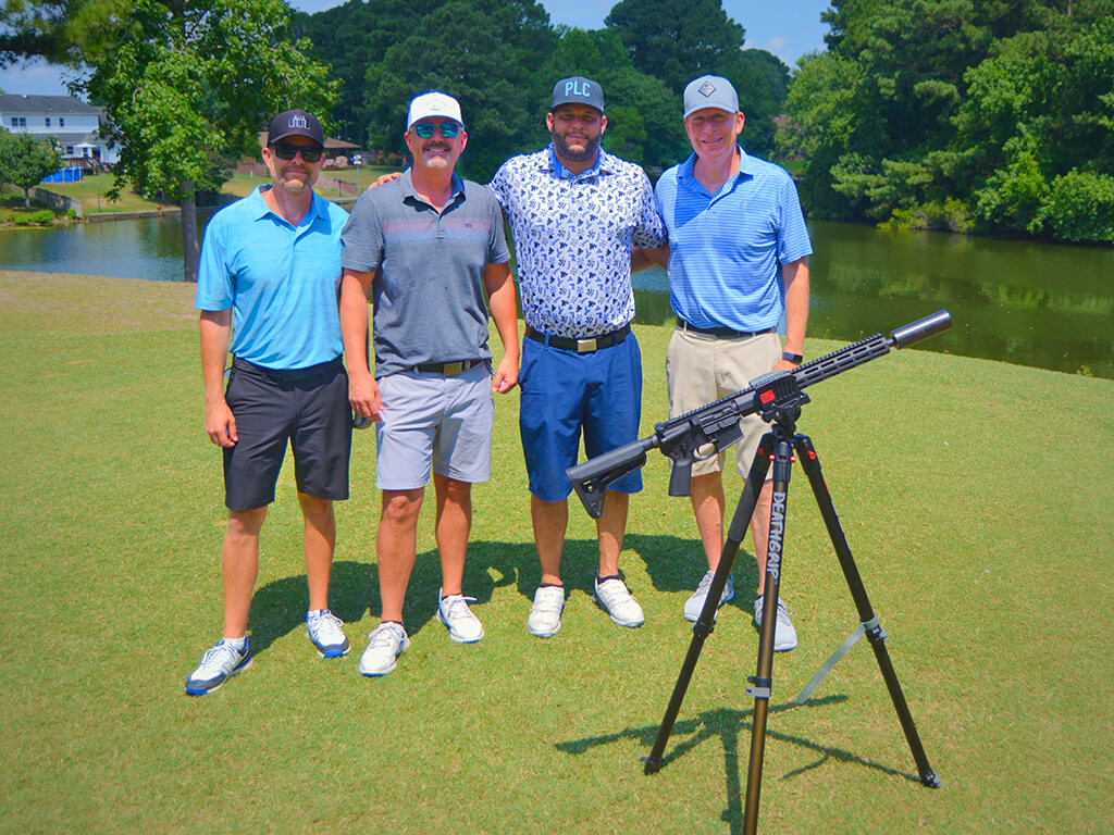 This was team 15. A group of four men standing with arms around each other, on the green by the water with trees behind them. The launcher is in front of them.