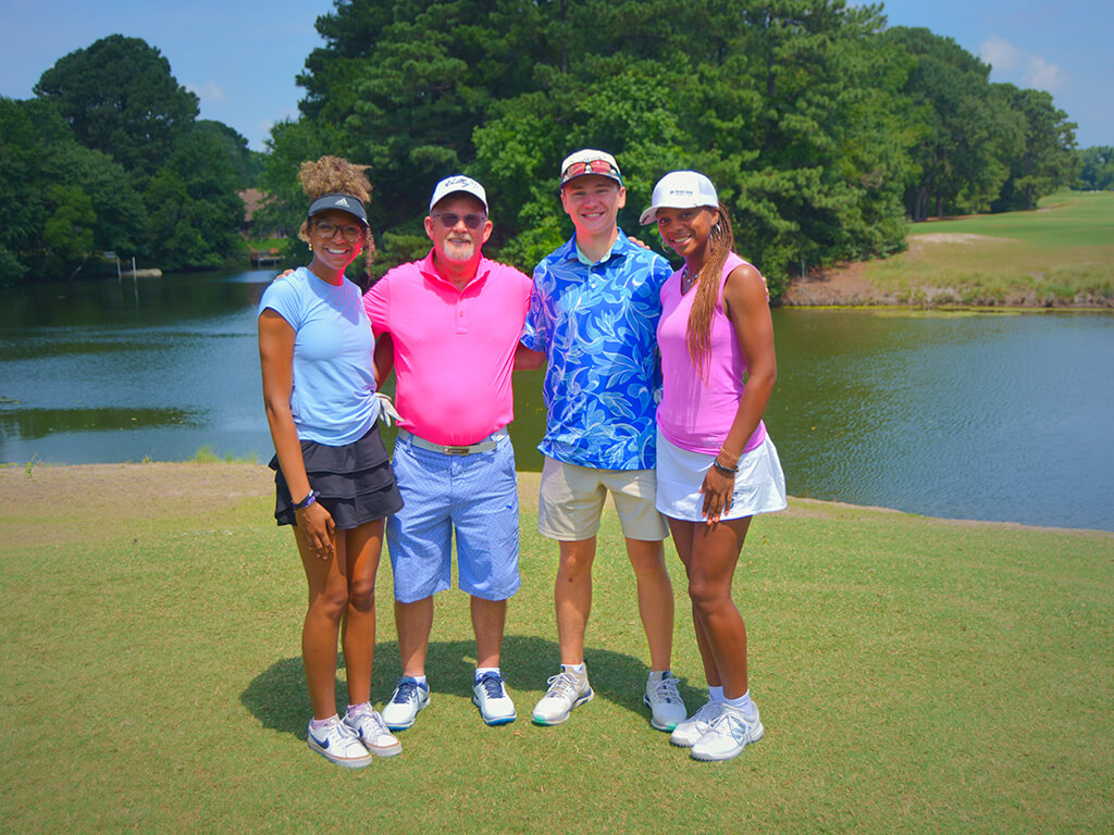 This was team 16. A group of two men and two women standing with arms around each other, on the green by the water with trees behind them.