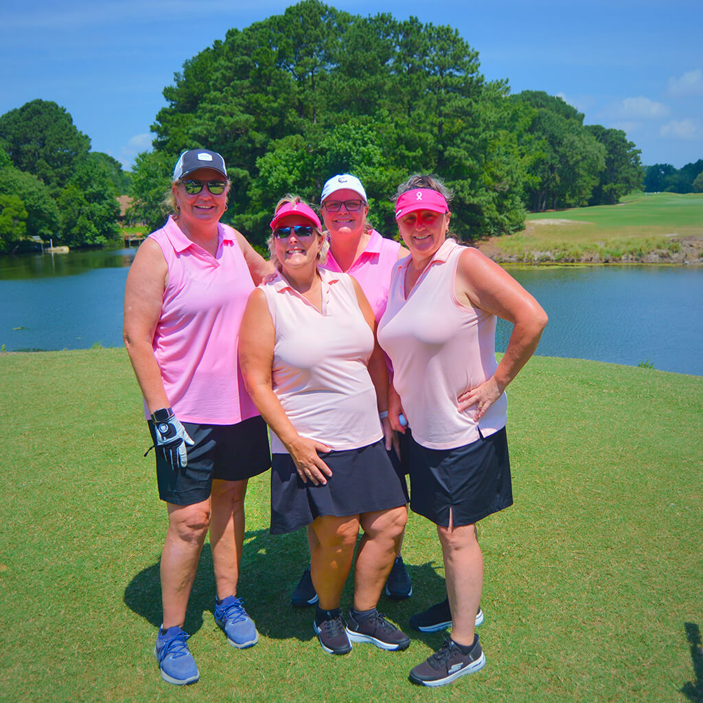 This was team 17. A group of four women standing close together with hands on hips, on the green by the water with trees behind them.
