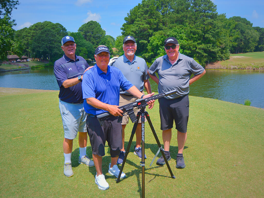 This was team 18. A group of four men standing on the green by the water with trees behind them. The launcher is in front of them. One of the men is holding the launcher.