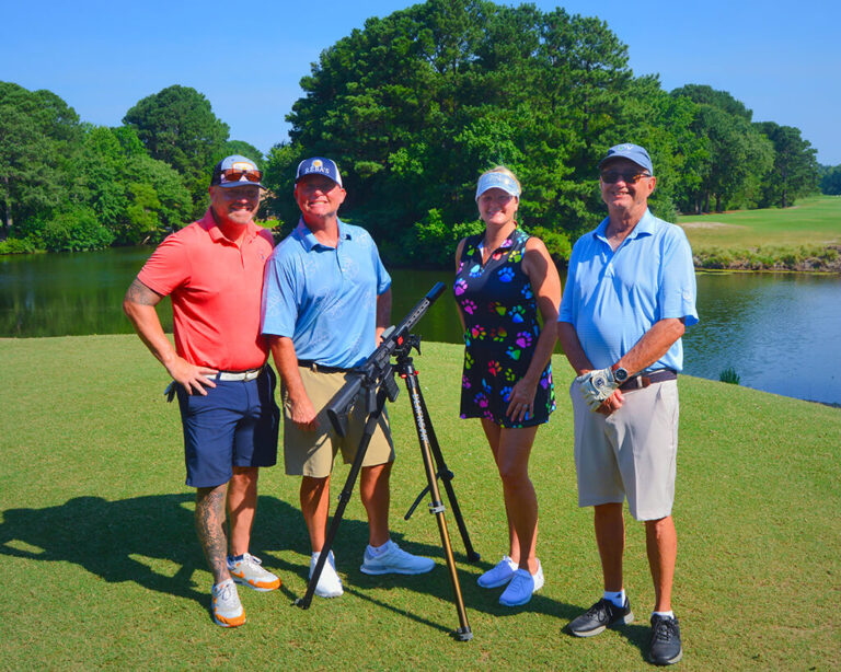 This was team 2. A group of four people gathered around the AR15 ball launcher. Three men and one women standing on the green by the water with trees behind them.