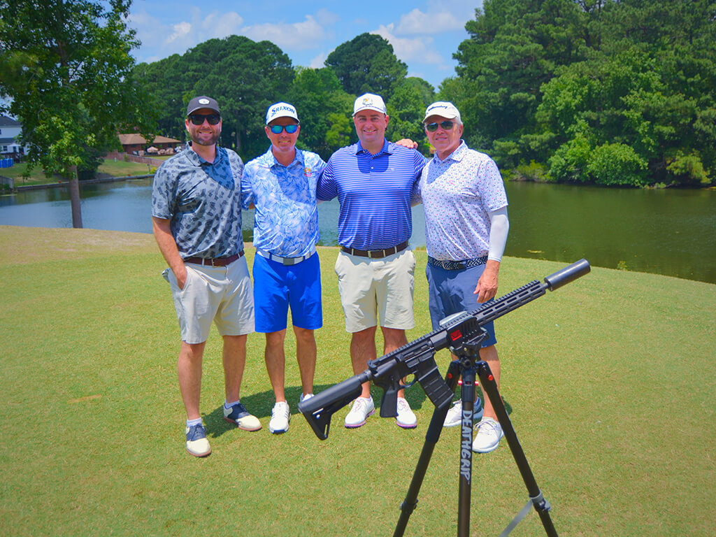This was team 21. A group of four men with arms on each others shoulders standing on the green by the water with trees behind them. The launcher is in front of them.