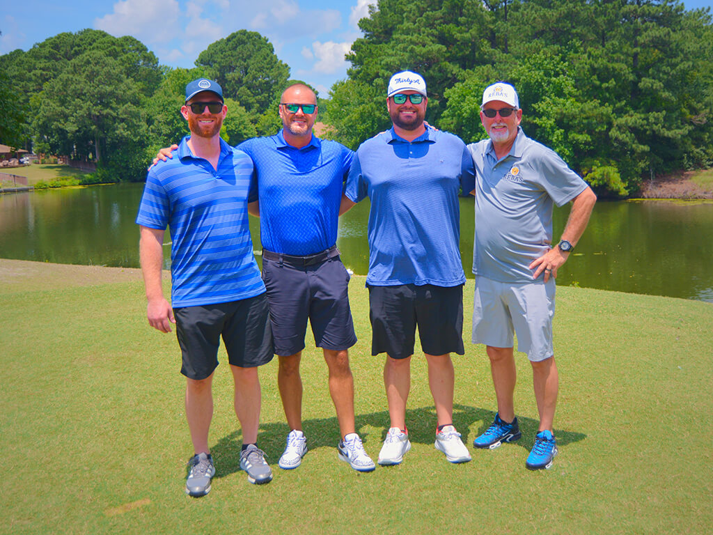 This was team 22. A group of four men with arms on each others shoulders standing on the green by the water with trees behind them.
