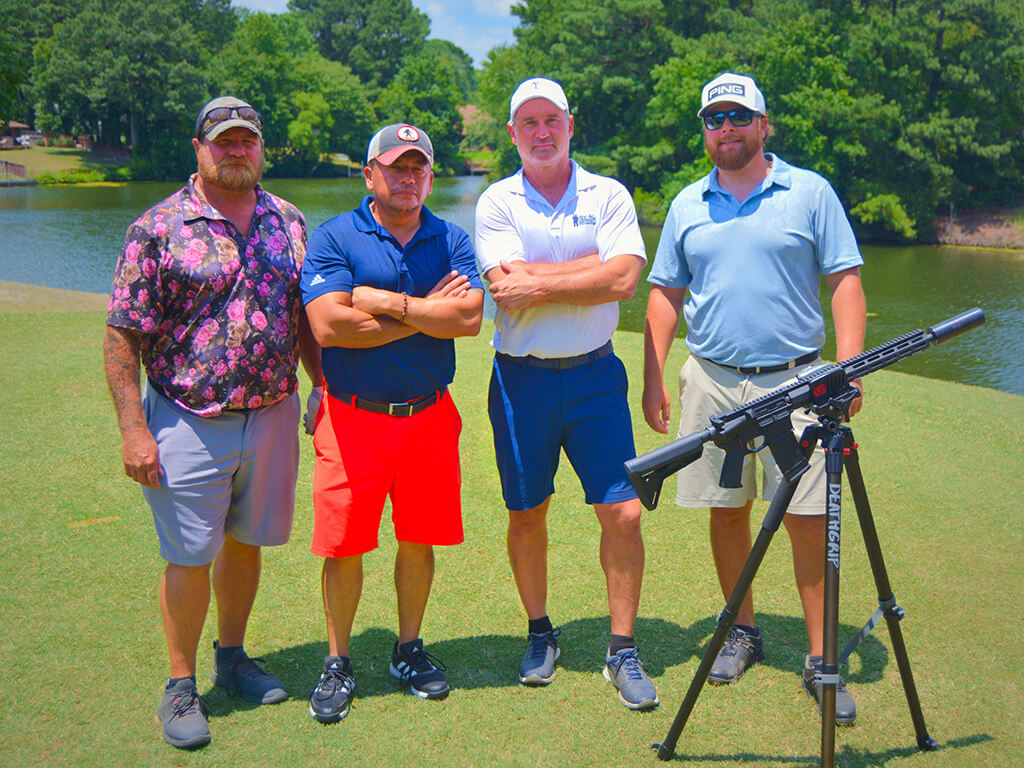 This was team 25. A group of four men standing on the green by the water with trees behind them. The launcher is in front of them.
