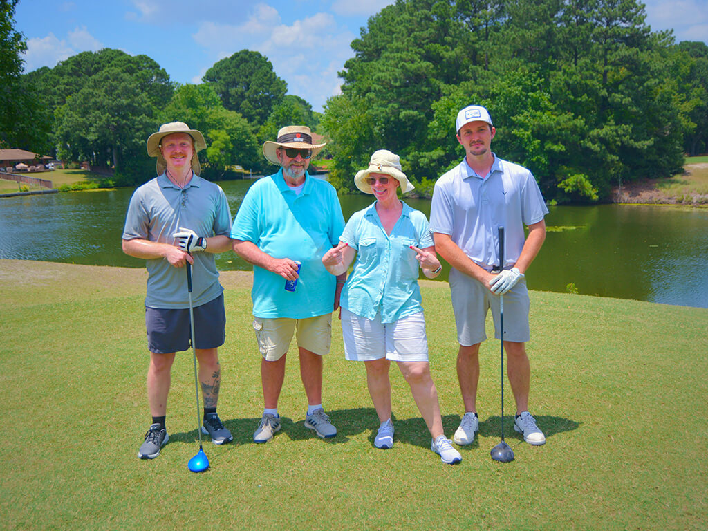 This was team 26. A group of three men and one woman standing on the green by the water with trees behind them. Two men are holding golf clubs