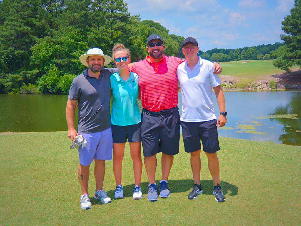 This was team 27. A group of three men and one woman with arms around each other standing on the green by the water with trees behind them.