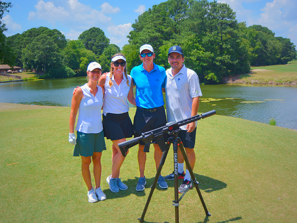 This was team 28. A group of two men and two women with arms around each other standing on the green by the water with trees behind them. The launcher is in front of them.
