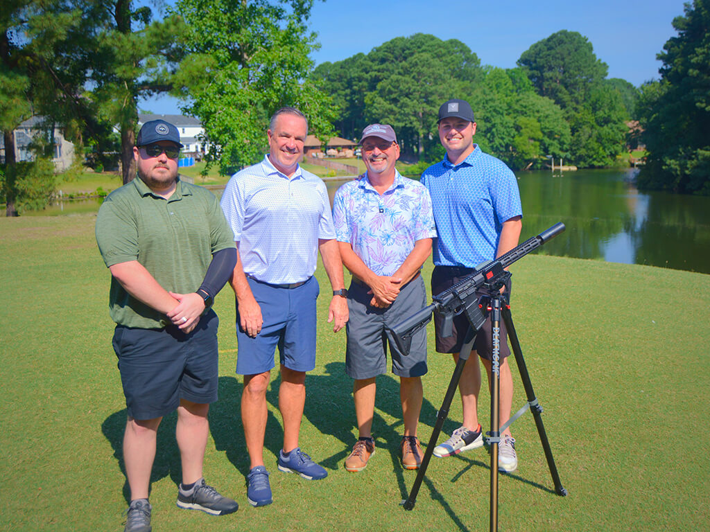 This was team 3. A group of four men gathered around the AR15 ball launcher standing on the green by the water with trees behind them.