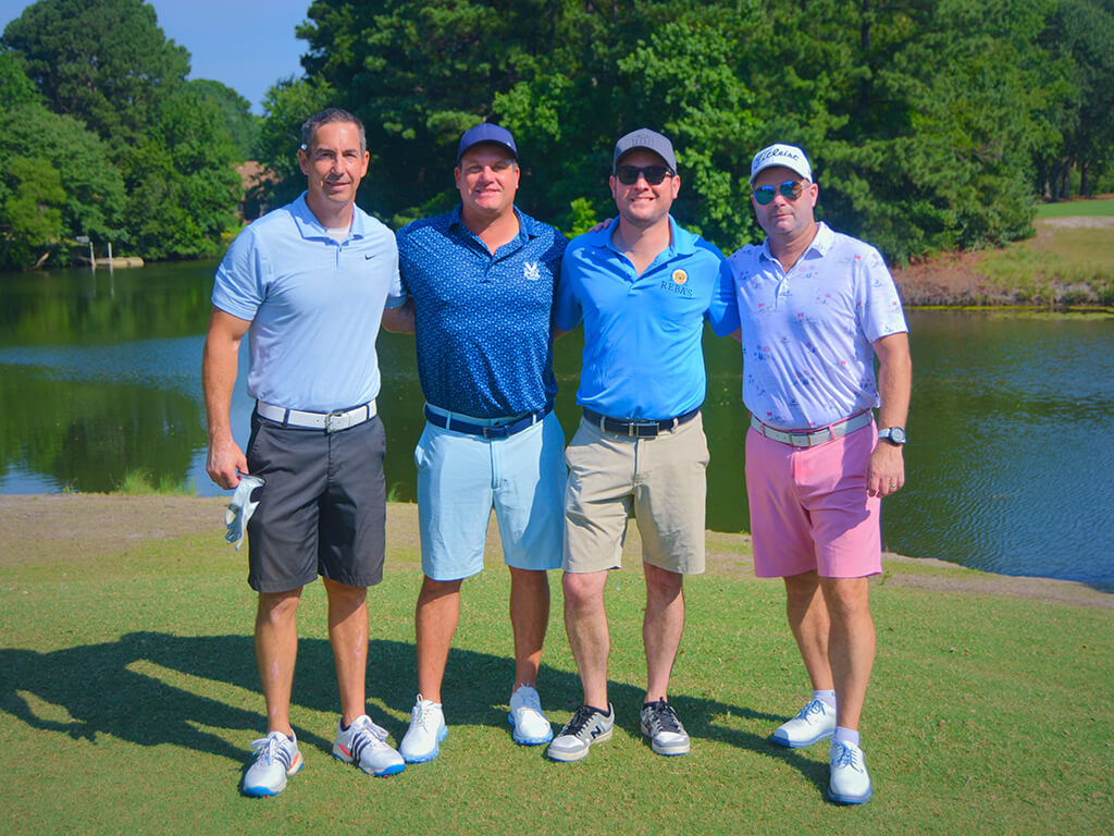 This was team 4. A group of four men with their arms around each other standing on the green by the water with trees behind them.