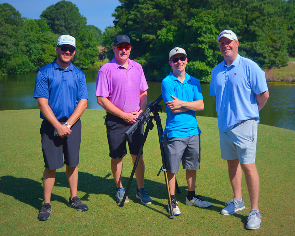 This was team 5. A group of four men with their hands crossed in front of them standing on the green by the water with trees behind them. The launcher is in between them.