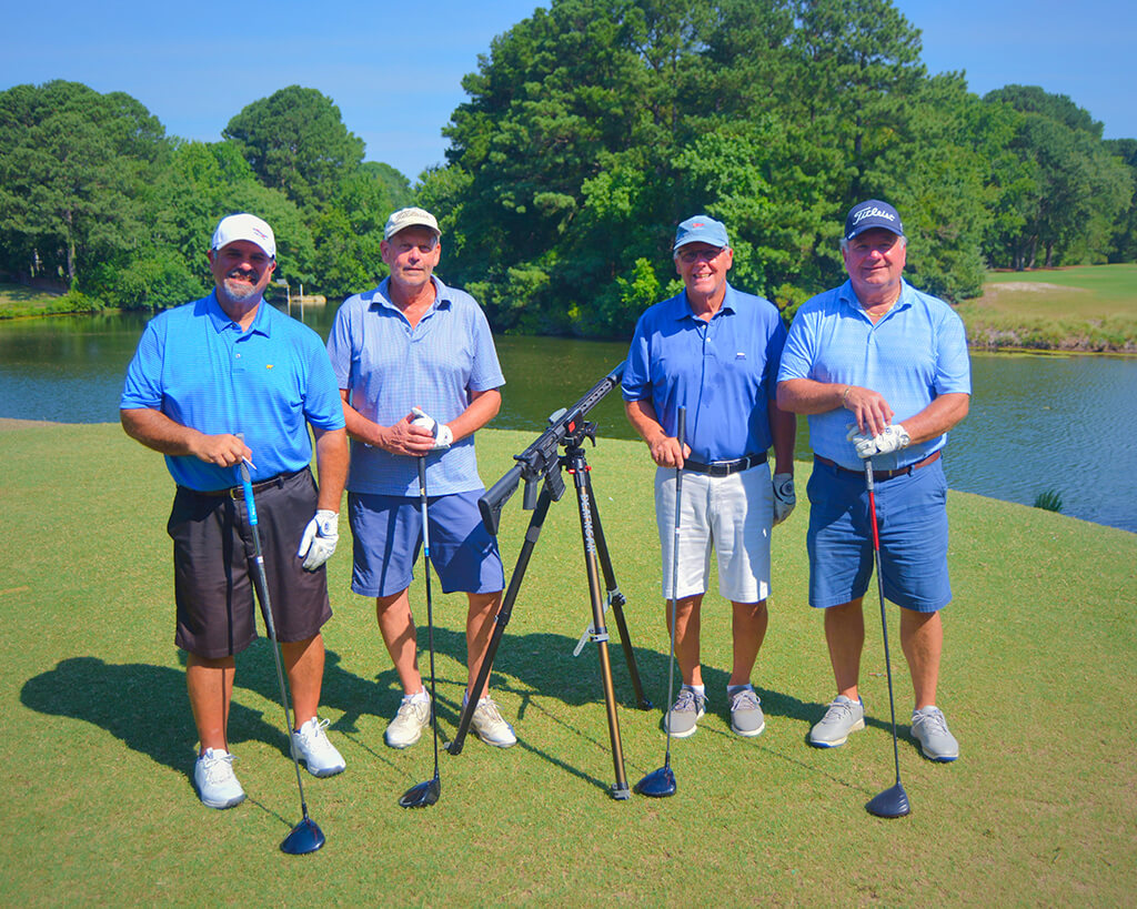 This was team 6. A group of four men with blue shirts standing on the green with their golf clubs by the water with trees behind them. The launcher is in between them.