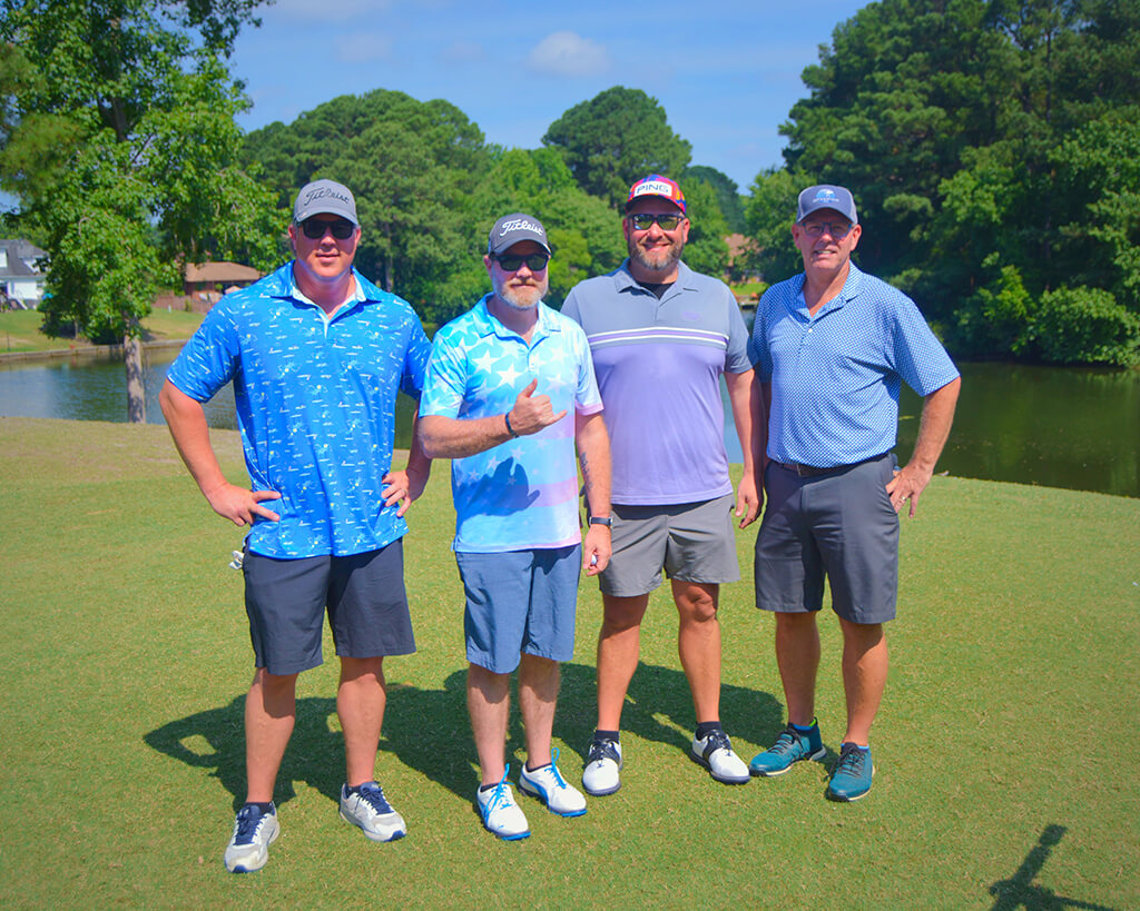 This was team 8. A group of four men standing on the green by the water with trees behind them.