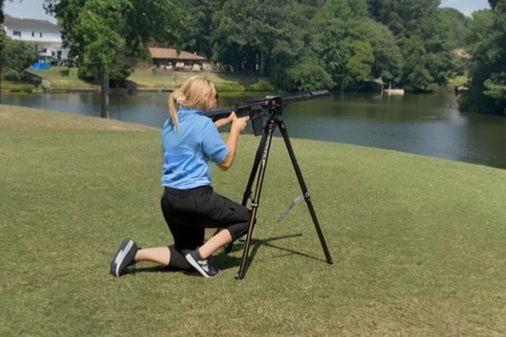 Meagan Goins is on one knee in blue shirt and black pants, shooting the AR15 golf ball launcher. the background includes the golf green, water, trees and houses.