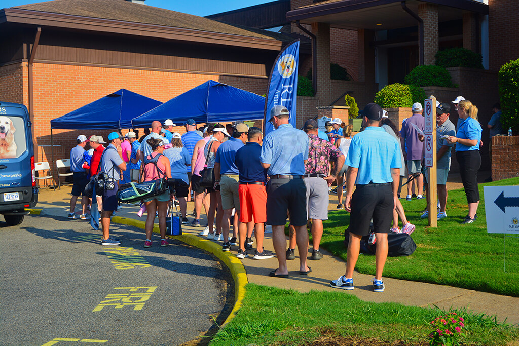 A crowd of golfers gathered in front of the country club waiting in line for registration. There are two blue tents and a Rebas flag and van by the tents.