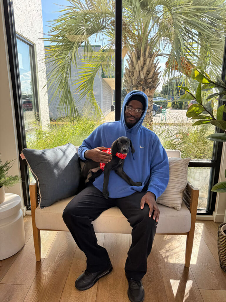 One man in a blue hoodie holding a black puppy wearing a red harness.