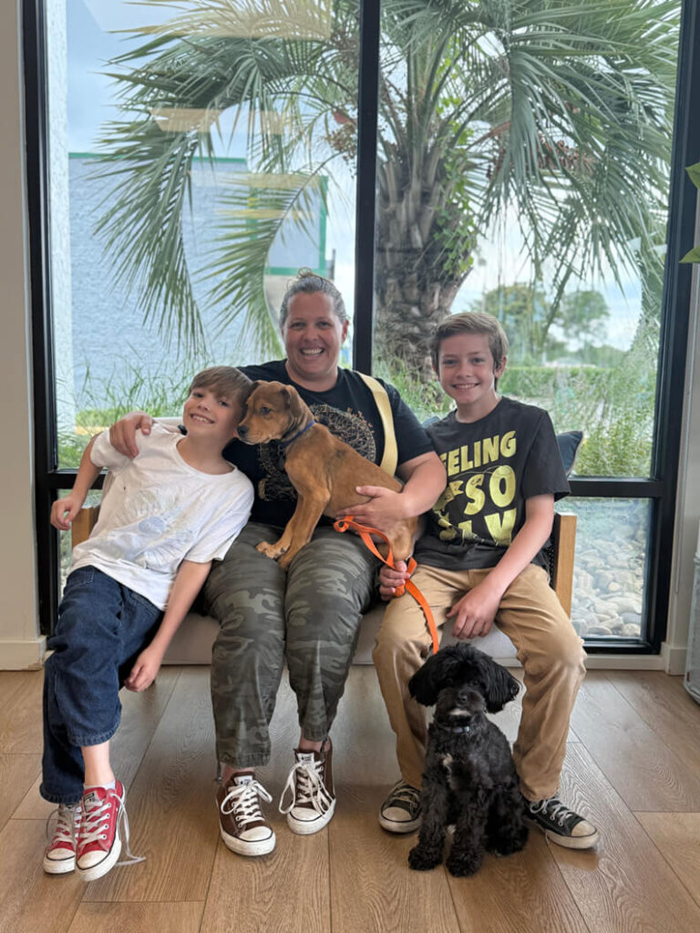 Mother with two kids holding a new brown puppy and a black dog is sitting on the floor.