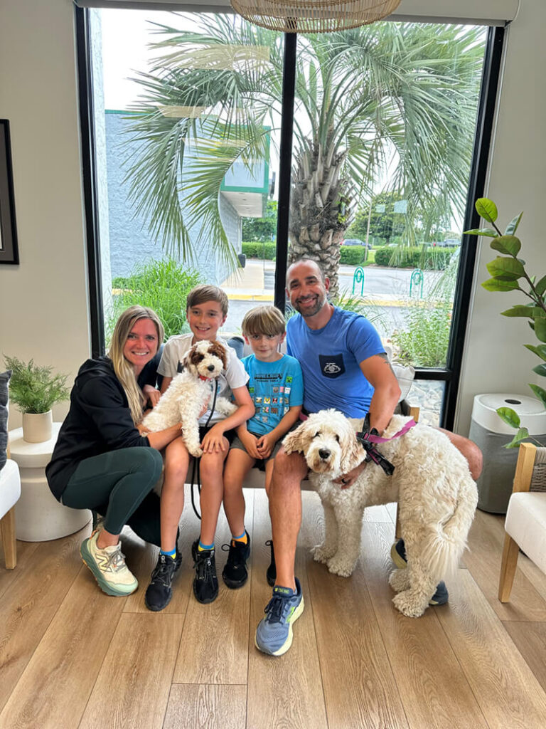 Family of four holding a new curly white puppy with a brown head. Their existing white doodle is pictured with the adoption.
