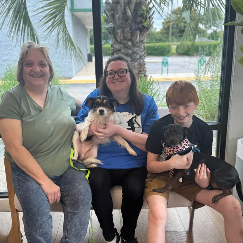 Family of three with 2 medium size dogs on their laps. One is white with a black head and the other is black and dark brown.