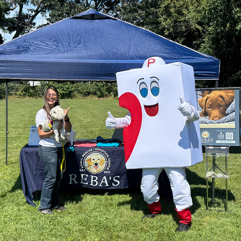 Volunteer holding white dog next to the Mr Puzzle Piece in front of Rebas booth