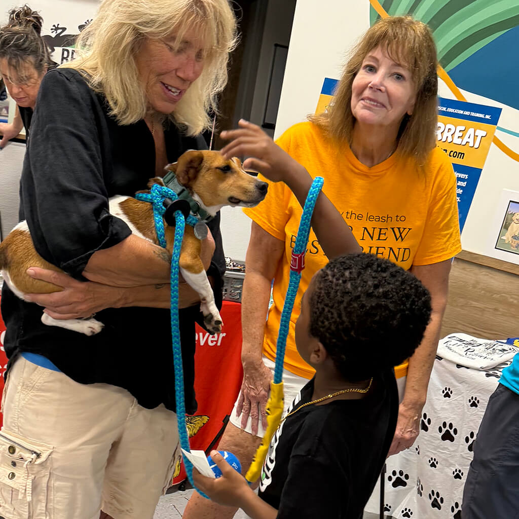 Little boy is reaching up to pet the brown dog's head while the lady holds him.