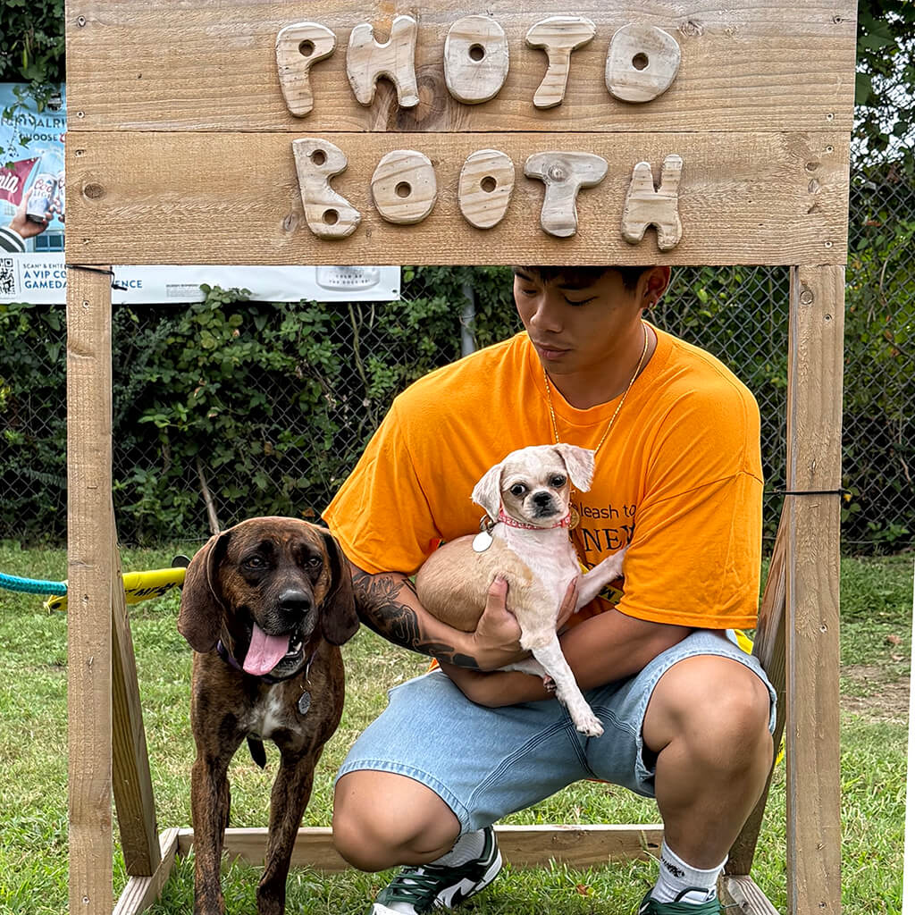 Young man in orange shirt is holding the little tan dog behind the wooden Photo Booth. The large brindle dog is standing next to them with his tongue out.