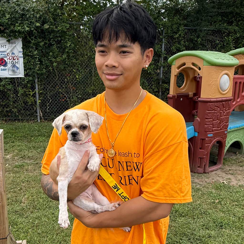 Young man in orange shirt holding the little tan dog.