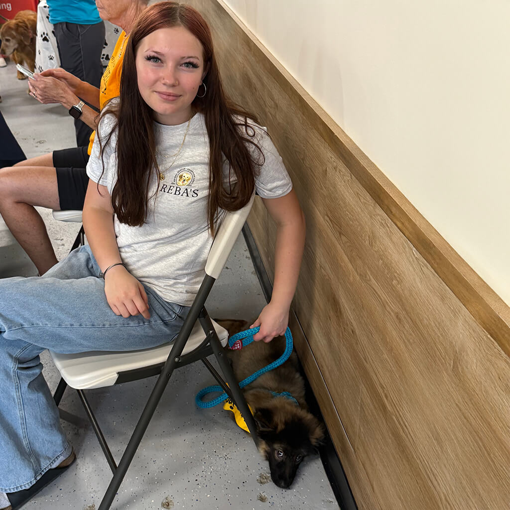 Lady with long hair and gray Reba's t-shirt sits in chair with dog relaxing behind chair.